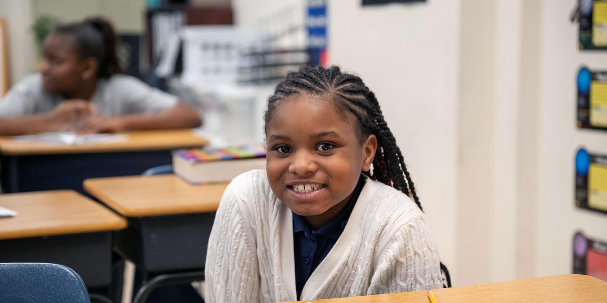 Smiling student with braids and a white cardigan seated at a classroom desk.