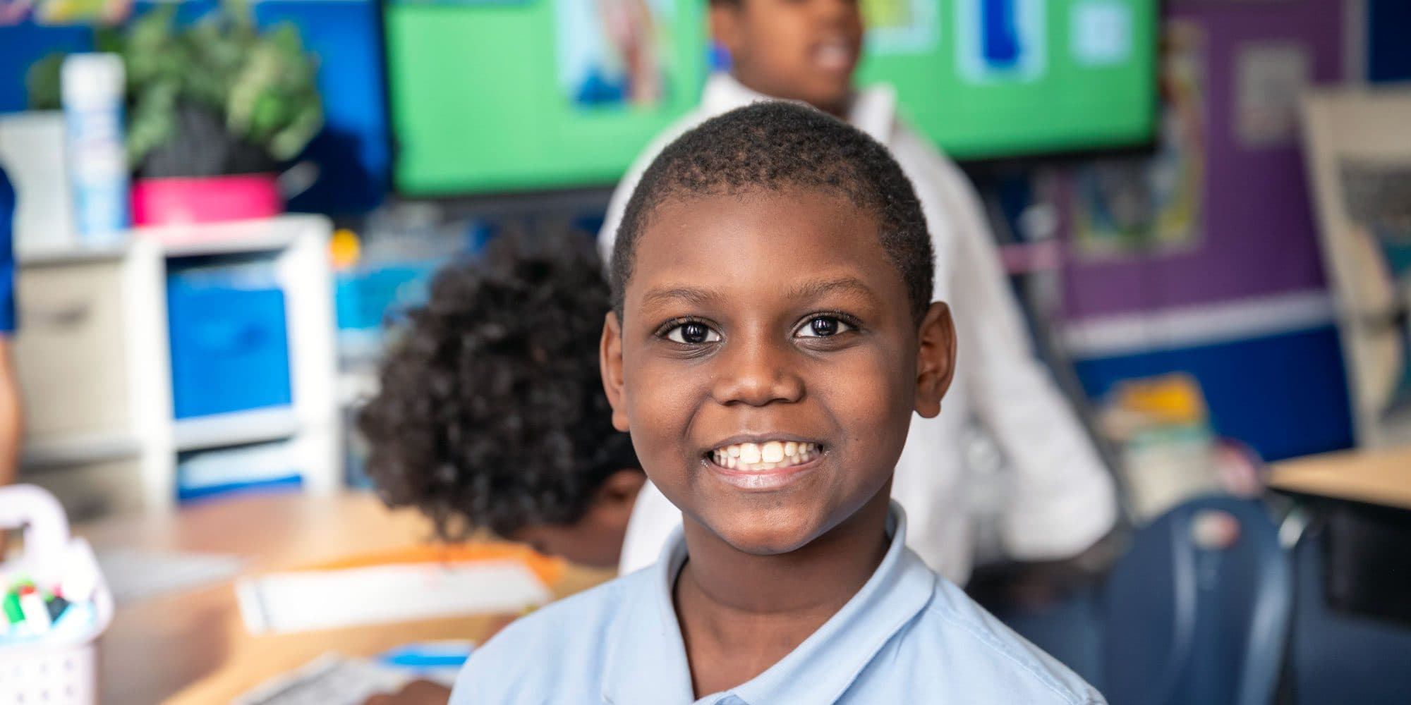 Smiling student in a light blue polo shirt in a busy classroom.