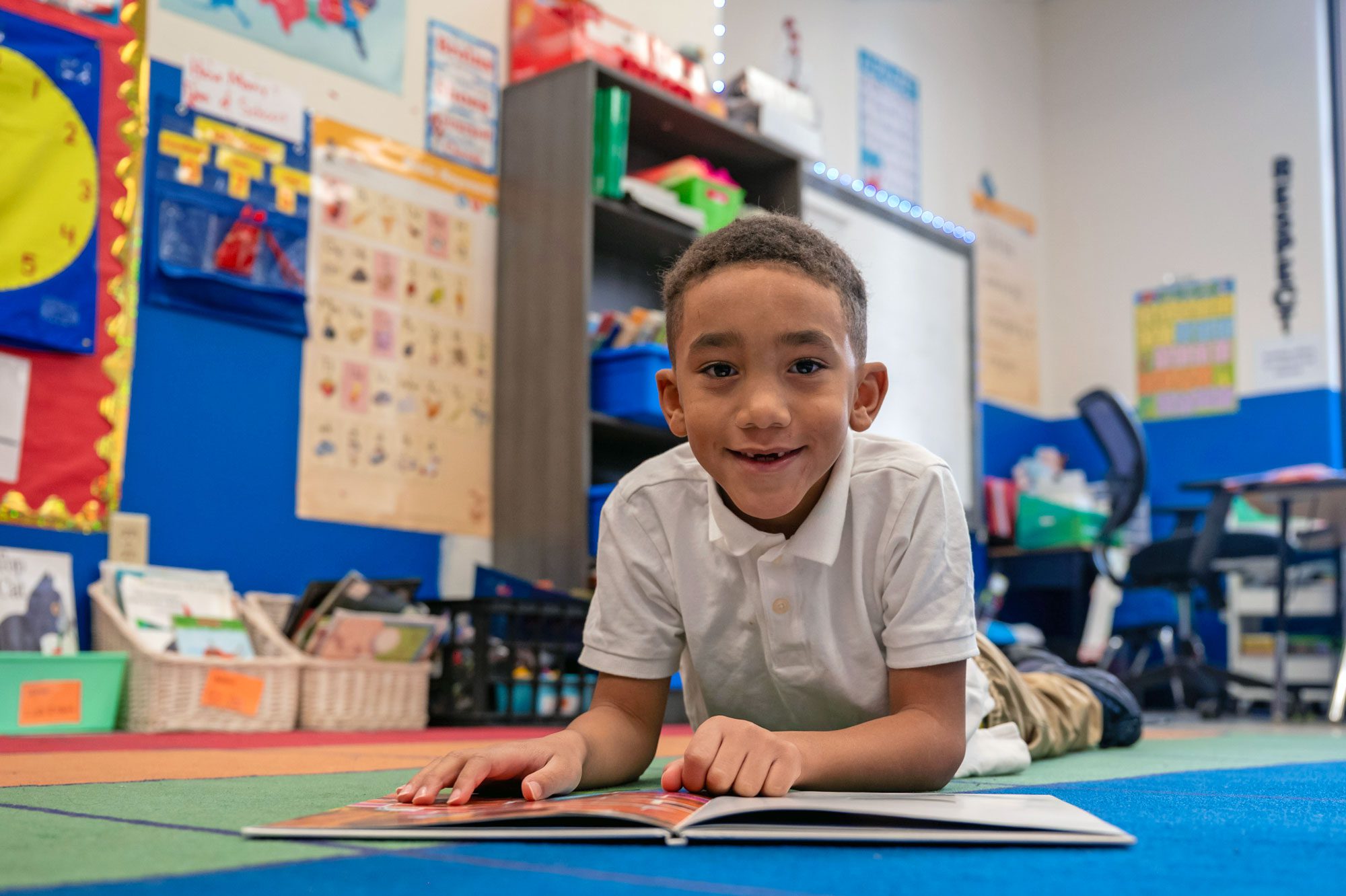 Young student smiling while reading a book on a classroom rug.